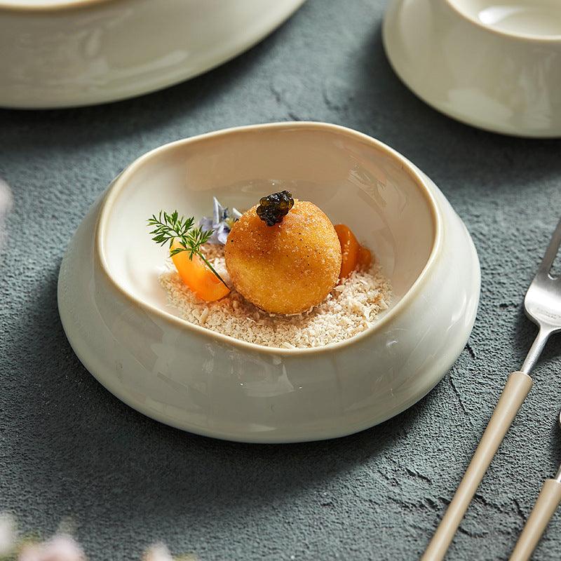 A gourmet dish presented on a ceramic plate with an irregular shape. The plate is beige in color, and there is a decorative element and a piece of food, possibly a dessert, presented on the plate.
