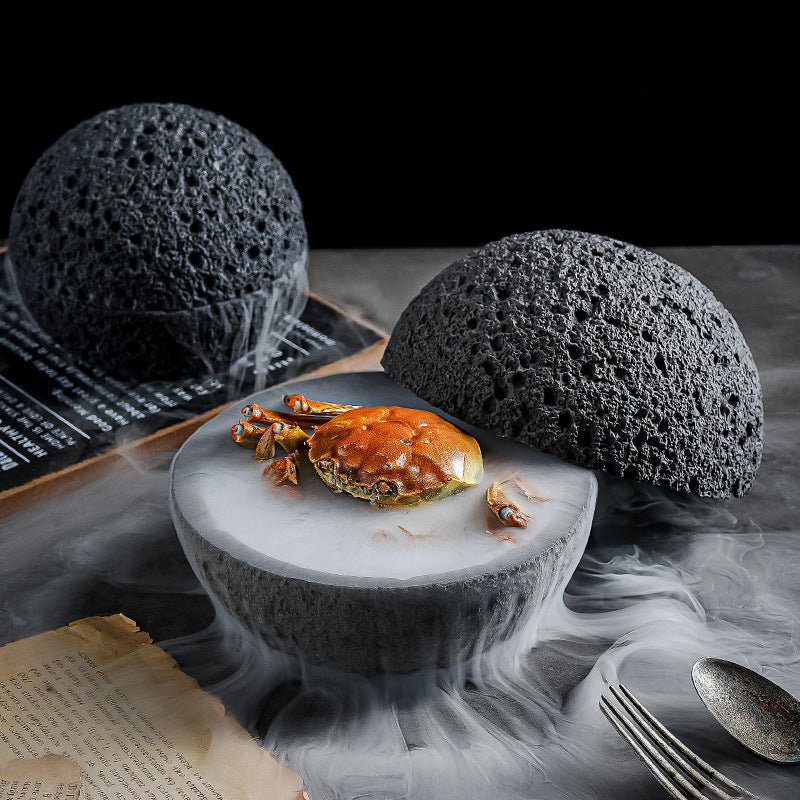 Close-up of a cooked crab centered in a split, volcanic-textured white bowl on a dark surface with theatrical white smoke.