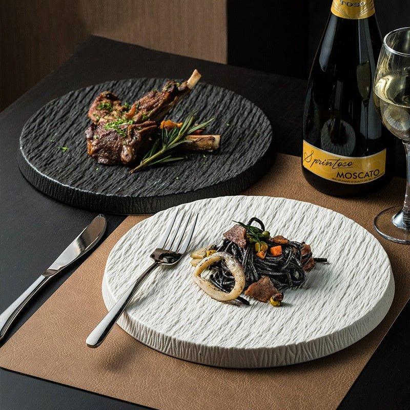 Two ceramic flat plates, one black and one white, with a rock pattern texture, displayed on a table with food items and a glass of wine in the background.