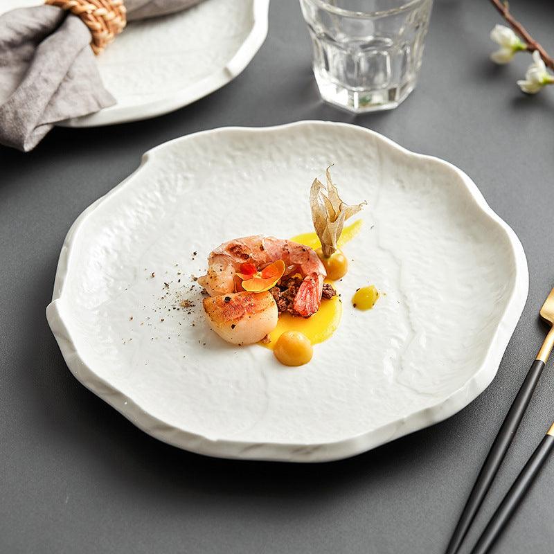 A white ceramic plate with a scalloped edge presented with a plated dish consisting of shrimp and garnishes on a gray surface, accompanied by a glass and a napkin.