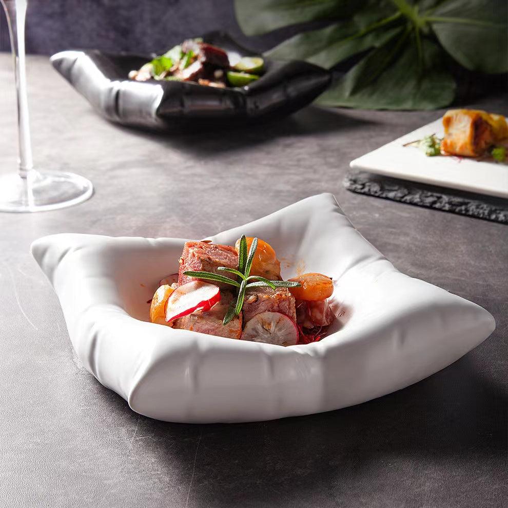 A cushion-shaped white ceramic plate with an irregular edge presented on a table, containing a meal consisting of meat and vegetables garnished with a herb.