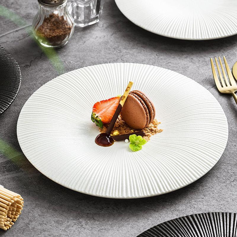 A white ceramic plate with a ridged edge design, displaying a plated dessert consisting of a chocolate mousse, strawberry, and a macaroon.
